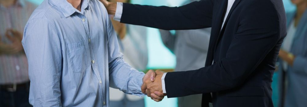 A business professional in a dark suit shaking hands with a casually dressed individual, with a blurred background of people clapping, indicating a recognition or congratulatory event for Offshore Development