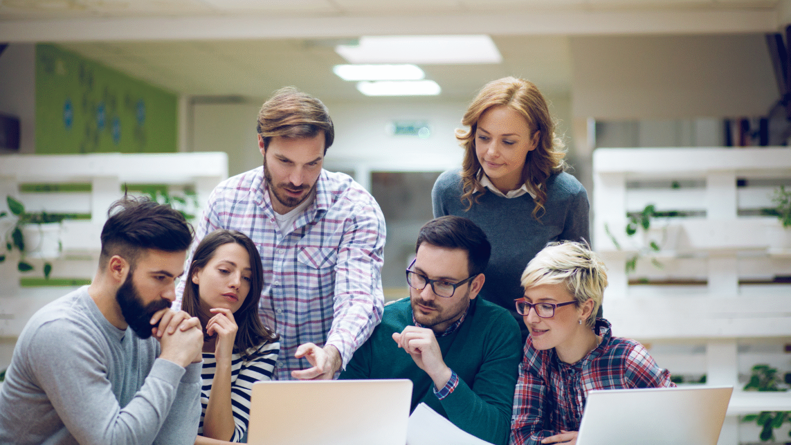A group of six young professionals collaborating around a laptop in a modern office setting. One man in a plaid shirt is pointing at the screen while others attentively listen and discuss. The diverse team appears engaged in brainstorming or troubleshooting, representing teamwork in software maintenance and ongoing system support.