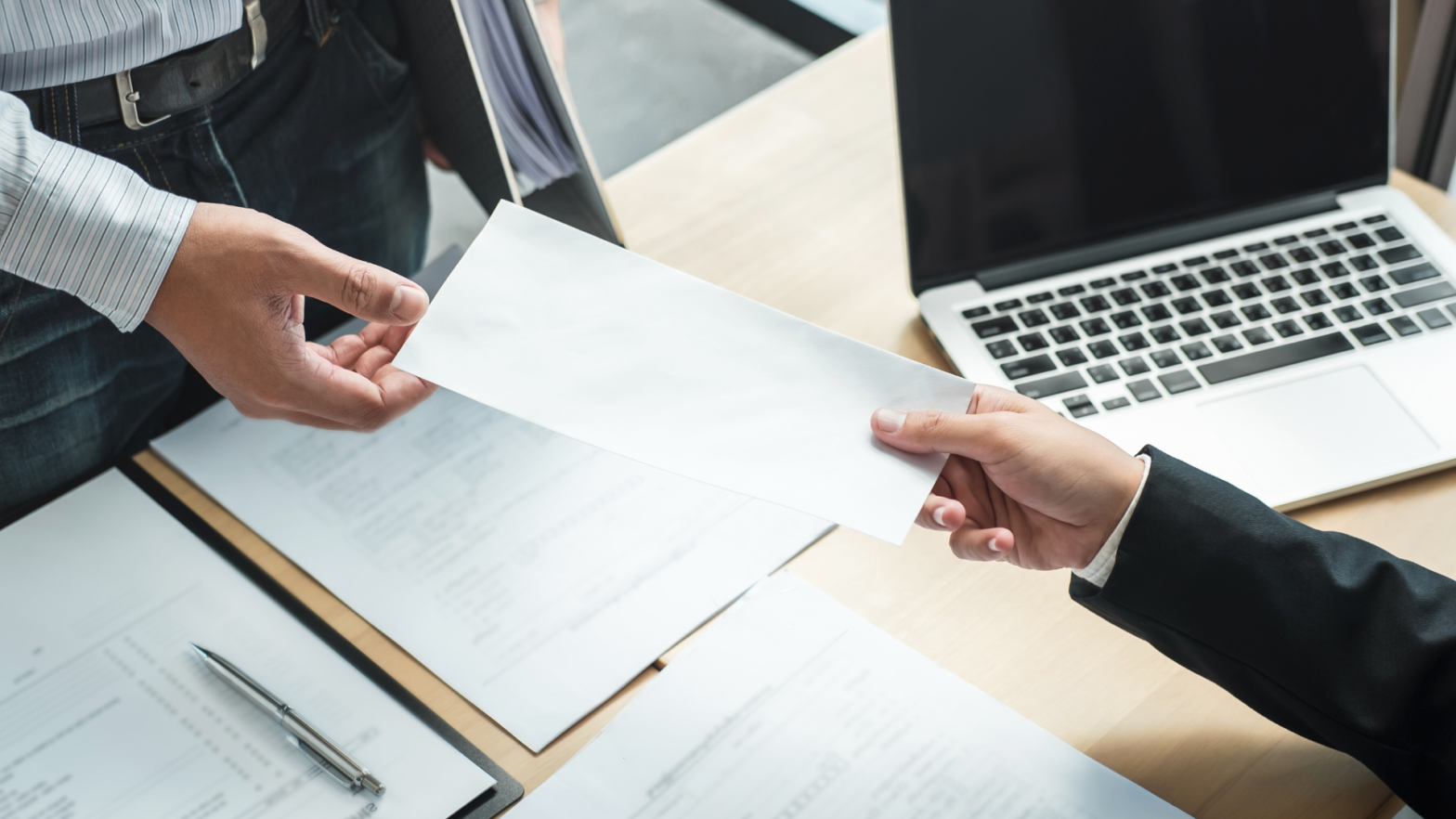 Employee handing over a white envelope, symbolizing a resignation letter, to a manager across a desk with documents and a laptop.